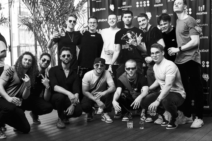 A black-and-white group photo of approximately 14 people posing together at what appears to be a tech or blockchain event, with sponsor logos visible in the background including 'blockchain', 'mimo', 'ig.store', and 'phabot'.
