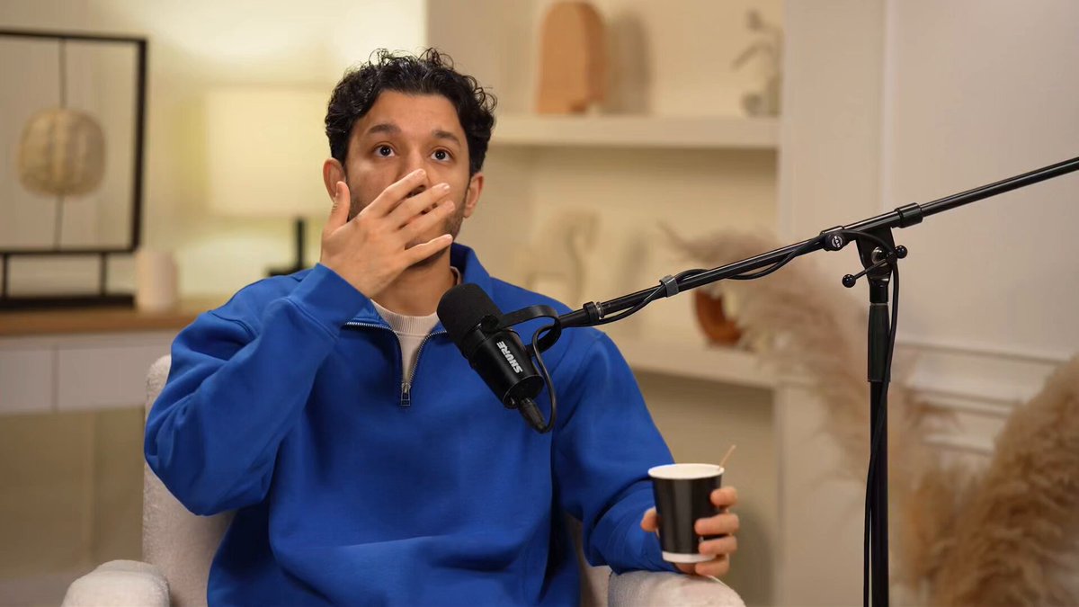 The image shows a man sitting down with his hand over his mouth while holding a cup of coffee. The background features a microphone setup, suggesting that he might be participating in an interview or speaking at an event. The man appears to be wearing a blue sweater, and there is a potted plant nearby.