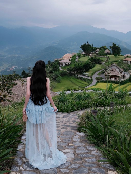 A woman in a flowing light blue dress stands on a stone pathway, gazing out over a serene mountain village with thatched-roof houses nestled among terraced green fields and misty peaks. The image beautifully captures the contemplative mood of the accompanying post, embodying that quiet moment of stillness and reverence for nature's breathtaking vastness.