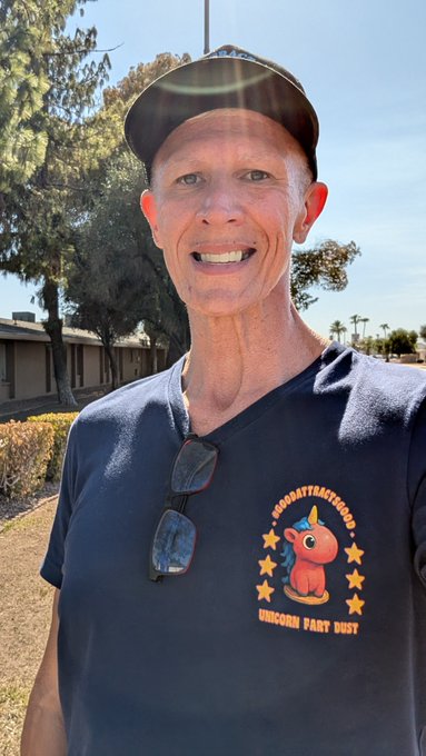 A smiling man wearing a cap and a navy 'AZOPATTYACTICGHOST Unicorn Party Guy' t-shirt with a cartoon unicorn poses outdoors with sunglasses hanging from his shirt.