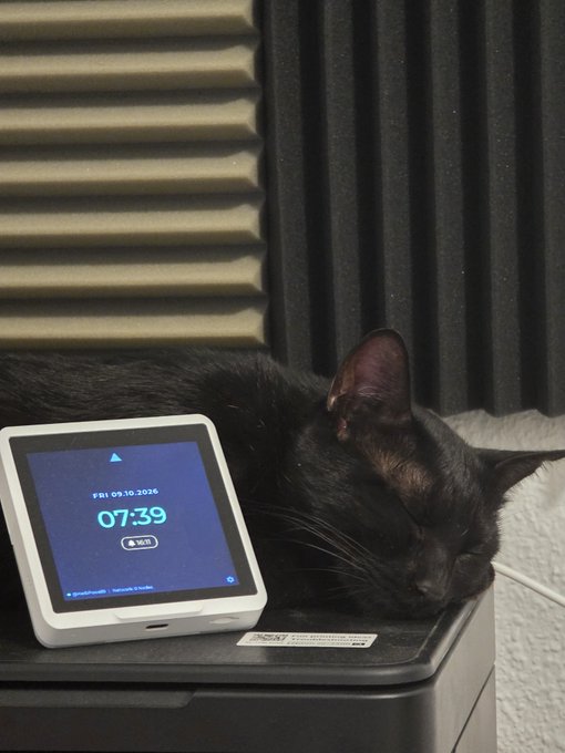 A black cat sleeps peacefully on top of a smart home display showing the time 07:39 and date FRI 09.10.2026, positioned in front of acoustic foam panels.