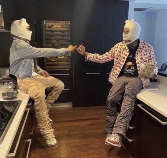 Two people wearing white ski masks sit on kitchen countertops facing each other while exchanging what appears to be a small object between their fingers.