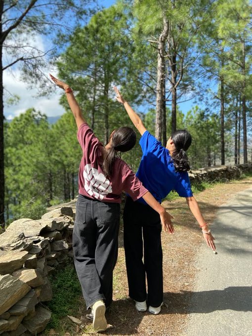 Two young women stand with their backs to the camera on a paved path in a forested area, raising their arms in playful poses next to a low stone wall.