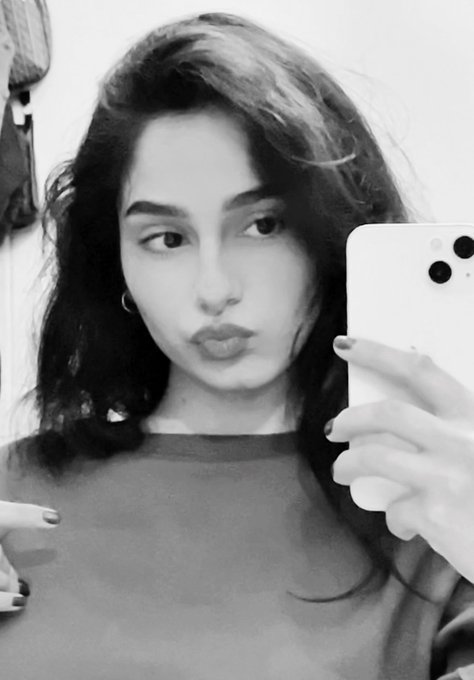 Black-and-white mirror selfie of a dark-haired woman wearing a gray t-shirt and holding a smartphone.
