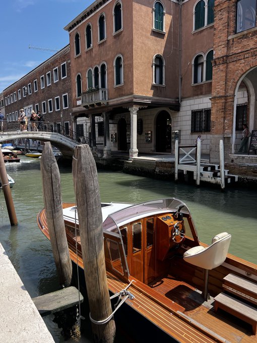 A scenic Venice canal view showing a polished wooden water taxi docked at wooden mooring posts, with historic brick buildings, a stone footbridge with pedestrians, and a clear blue sky in the background.
