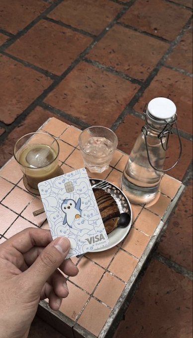 A hand holds a VISA card featuring a penguin character named Jerry, displayed over a tiled table with two glasses and a water bottle in the background.