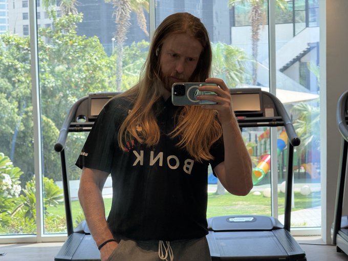 A gym selfie taken by a long-haired young man wearing a black 'BONK' t-shirt in front of a treadmill.