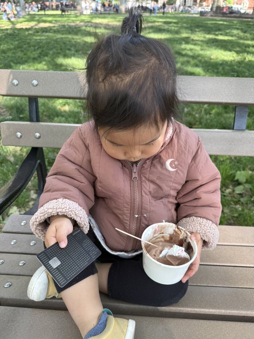 The image shows a young child sitting on a park bench, holding an ice cream cone with chocolate sauce and whipped cream. A cell phone can be seen in her hand as well. The photo also contains text that reads "She's buying the dip @coinbase". This suggests that the child is likely purchasing some ice cream or another treat from a nearby store using a mobile payment method, such as a credit card or digital wallet app like PayPal.

The scene takes place in an outdoor setting with grass and trees visible around the park bench where the child is seated.