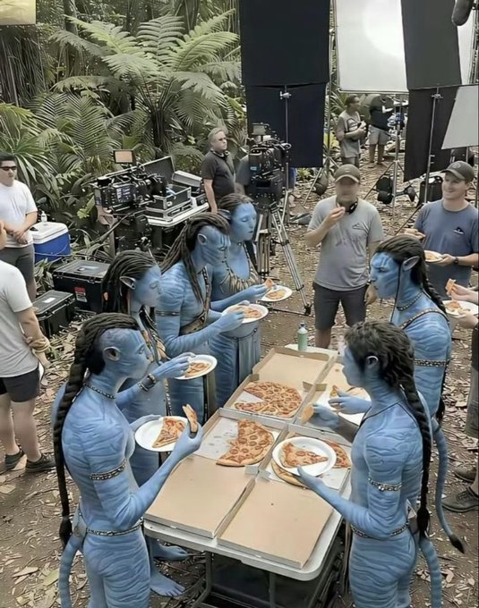 Behind-the-scenes photo from an Avatar film set showing several Na'vi characters in full blue body paint and makeup eating pizza around a table in a forest environment with film crew and equipment visible.