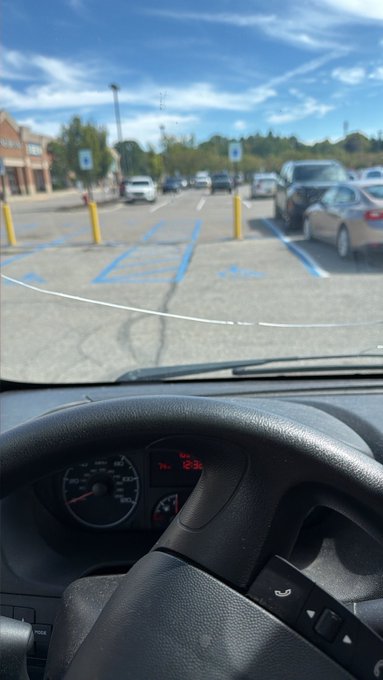 A view from inside a car looking out through the windshield at a sunny parking lot with blue-painted accessible parking spaces and yellow bollards.