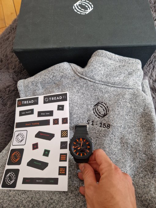 The image shows Tread Fi merchandise, including a grey sweater with the "S1-158" designation, a black G-shock watch with orange accents, and various stickers featuring trading-related graphics and phrases like "Tread Fi," "Take Profit," and "Stop Loss." This aligns with the accompanying post's excitement about receiving the merch.
