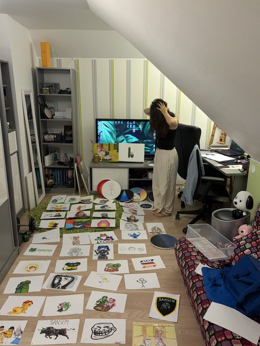 A woman stands in an attic room with a slanted ceiling, looking at a computer monitor displaying "CERES ELAR ZERO," while numerous colorful drawings are spread across the floor.