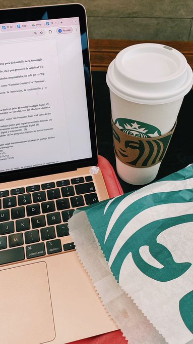 A study setup featuring a rose gold laptop displaying a document, a white Starbucks coffee cup, and a green Starbucks paper bag resting on a wooden surface.