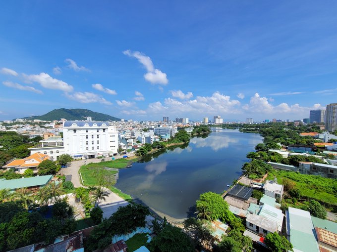 An aerial photograph captures a scenic cityscape featuring a calm lake reflecting the sky, surrounded by buildings and lush green trees under a bright blue sky with scattered clouds.