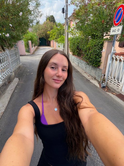 A young woman with long brown hair, wearing a black top with a purple bikini strap showing, takes a selfie on a quaint residential street lined with greenery. A "no parking" sign and an arrow sign pointing left are visible in the upper right corner, complementing her cheerful "GM have the best day!" morning greeting.