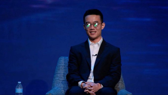 A man wearing glasses, a white shirt, and dark blazer sits smiling on a stage chair with a water bottle beside him, likely a speaker or builder being referenced in the discussion about Pumpfun's integration of Hyperliquid perps.