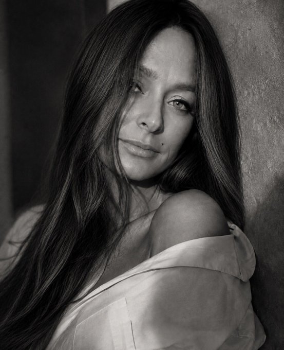 A black and white portrait of a woman with long dark hair looking pensively over her bare shoulder against a textured wall.
