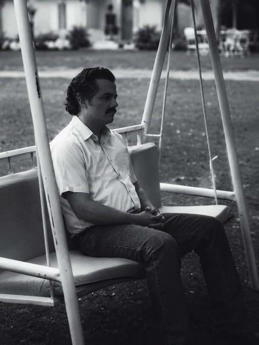 A black and white photograph of a man with a mustache sitting on a white swing in a garden, wearing a short-sleeved button-up shirt and jeans, looking off to the side.