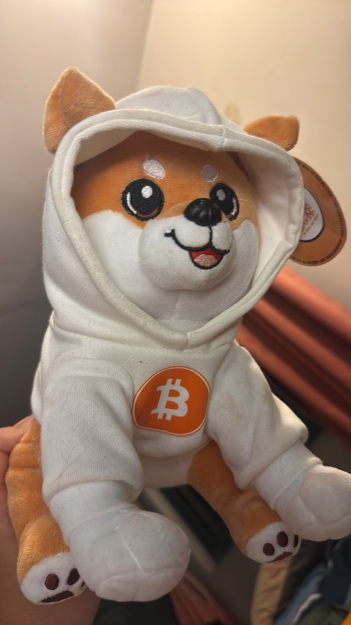 A plush Shiba Inu toy wearing a white hoodie with an orange Bitcoin logo on the chest, appearing to be a Bitcoin-themed mascot.