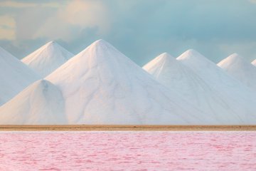 A landscape with piles of white material (possibly salt) shaped into pyramids against a cloudy sky, with a pink/red colored body of water in the foreground.