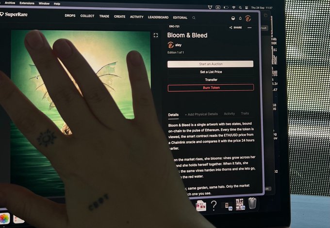 A hand with a small tattoo is held up in front of a computer screen showing the SuperRare NFT marketplace with a listing for an artwork titled 'Bloom & Bleed'.