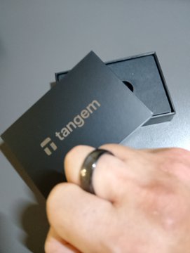 A close-up of a hand holding a small black box with the 'Tangem' logo, appearing to be a crypto hardware wallet packaging.