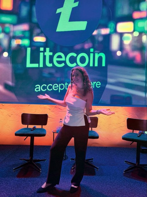 A woman in a white strapless top and dark pants stands on a stage in front of a large Litecoin logo with her arms outstretched in an enthusiastic, welcoming pose. The "#litecoin" hashtag aligns with the event's branding, as the backdrop prominently promotes Litecoin adoption with the partially visible slogan "acceptance."