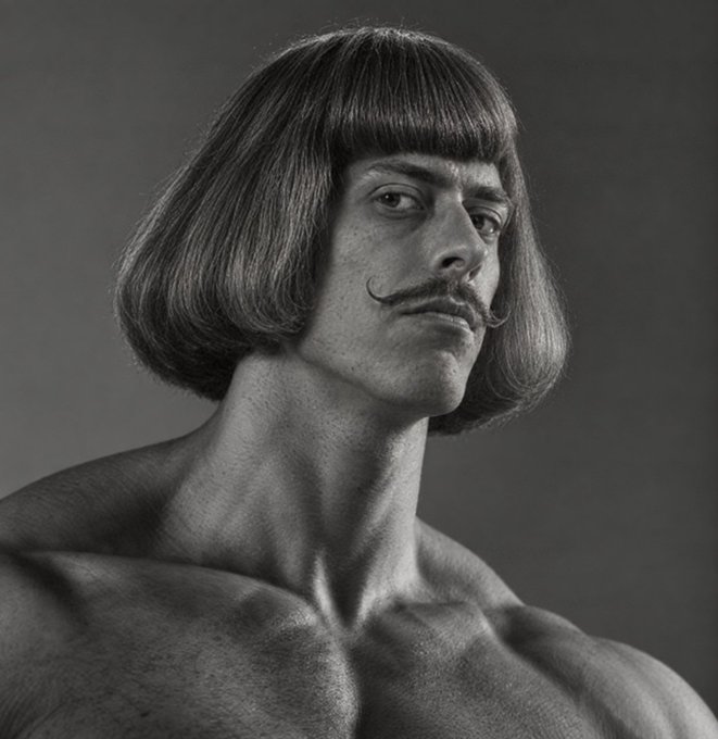 Black and white portrait of a shirtless muscular man with a distinctive bob haircut and curled handlebar mustache against a dark background.