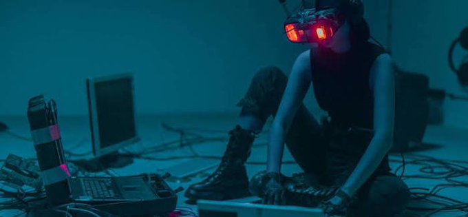 A person wearing red-lensed goggles sitting on the floor surrounded by electronic equipment and cables in a dimly lit, blue-toned room.