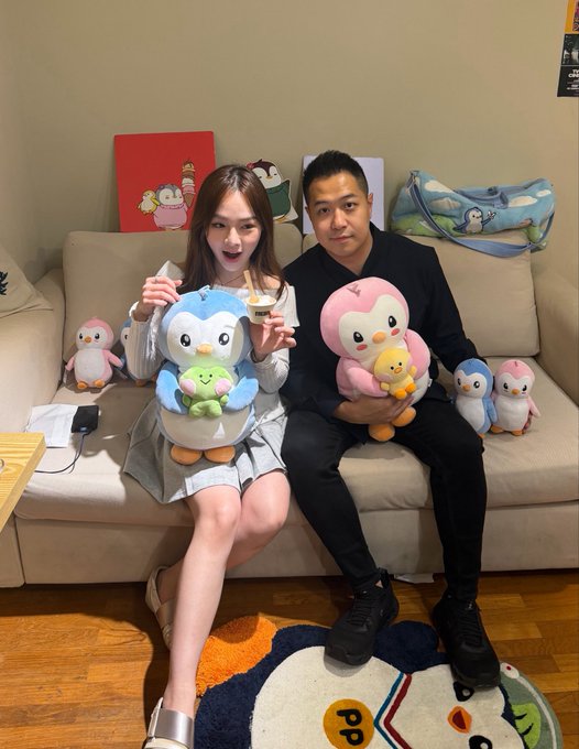 A man and woman sit on a couch holding plush toys of Pudgy Penguins characters, with additional penguin plushies and branded artwork visible around them, at what appears to be a Pudgy-themed social gathering. The setup reflects the post's reference to a "Pudgy Party," showcasing the brand's penguin mascot merchandise as they pose for a photo together.