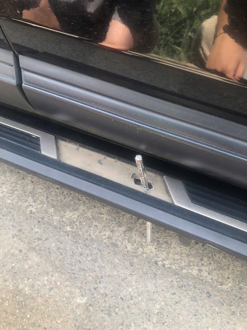 The image shows a black car parked on a street with its side door open. The car's silver bumper is visible as well. There are two people standing next to the car, one of them holding their hand out towards the car. A small metal object can be seen near the bottom right corner of the photo.