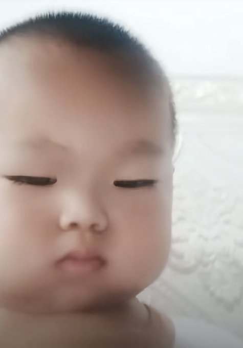 A close-up photo of a baby with half-closed eyes and a neutral expression, set against a soft white background.