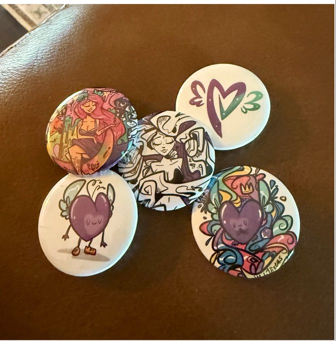 Five circular pins or badges are arranged on a wooden surface, featuring colorful heart illustrations, cartoon characters, and a black-and-white illustrated figure.