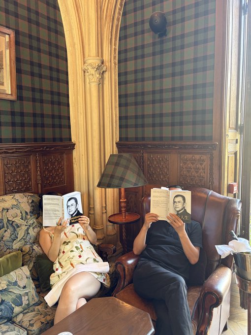 Two people sit in a room with green plaid wallpaper and wood paneling, each holding a book featuring a portrait of the same man and Chinese text, obscuring their faces.