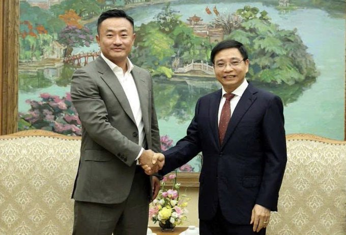 The image shows two men shaking hands, dressed in suits with one wearing a gray suit and the other wearing a black suit. They are standing next to each other near a couch, which has a painting of a lake behind them. The background features an oil painting depicting a landscape scene, adding an artistic touch to the setting.