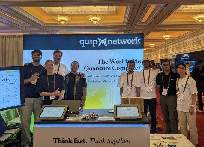 A group of people pose at a booth displaying 'qup network' signage, with text reading 'The Worldwide Quantum Computer' and 'Think fast. Think together.' on the front of the booth.