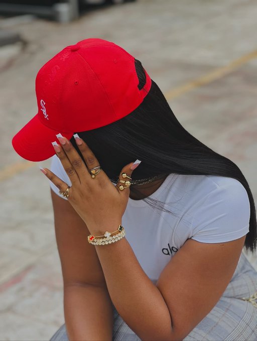 A woman in a red "South" cap and white "alo" t-shirt shields her face with her manicured hand, surrounded by gold rings and bracelets, embodying a moment of quiet self-reflection that aligns with the post's encouraging reminder for "builders" to acknowledge their progress.