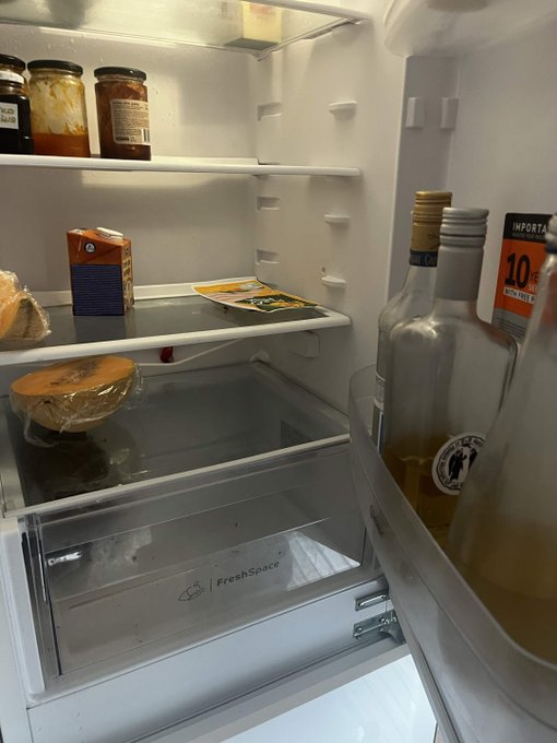 A nearly empty refrigerator containing only a few jars, a carton, some wrapped food items, and liquor bottles on the door shelf, with a FreshSpace crisper drawer visible.
