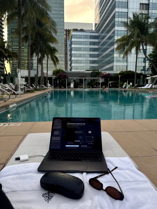 A workspace by a resort pool featuring a laptop displaying code, sunglasses, a mouse case, and a USB drive on a white towel, with tall buildings and palm trees in the background.