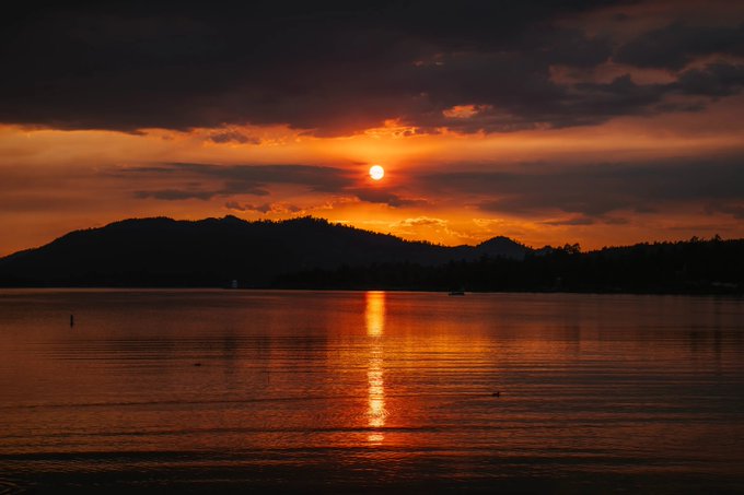 A serene sunset over a body of water, with the orange sun reflecting on the calm surface beneath a silhouette of mountains and dramatic clouds.