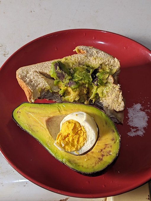 A red plate holds a halved avocado stuffed with a hard-boiled egg, served alongside a sandwich filled with avocado and other ingredients.