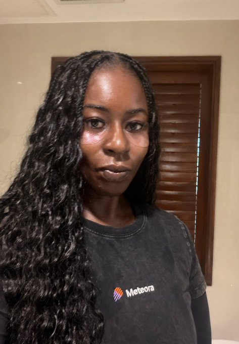 A woman with long dark braided hair wears a dark gray t-shirt with the 'Meteora' logo, posing for a selfie indoors with wooden blinds visible behind her.