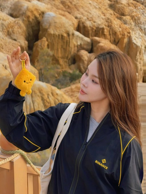 A young woman wearing a "BINANCE" jacket holds up a yellow plush keychain while standing against a rocky desert backdrop, visually reinforcing the post's reference to Binance VIP membership tiers and BNB holding thresholds.