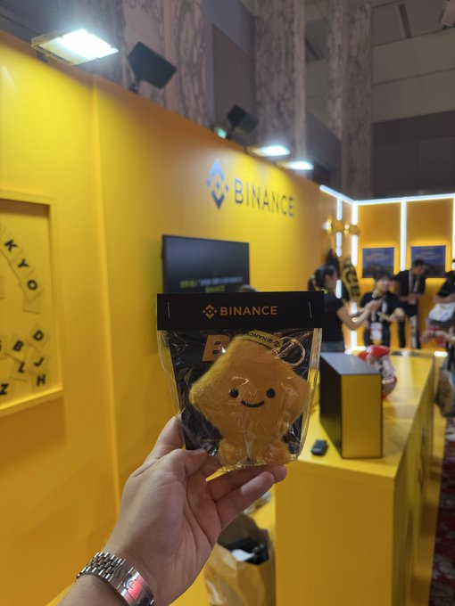 A first-person photo of a hand holding a packaged Binance-branded plush yellow character at what appears to be a Binance event booth, with a yellow Binance logo wall in the background.