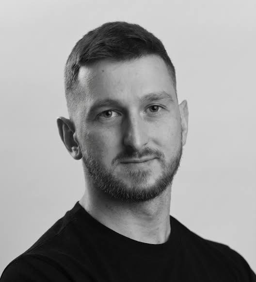 A black and white portrait of a man with a short beard wearing a black shirt, looking directly at the camera.