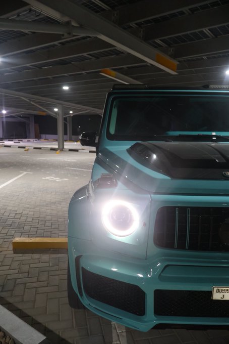 A turquoise Mercedes-Benz G-Class SUV with illuminated headlights parked in an underground parking garage, displaying a 'DUBAI J' license plate.