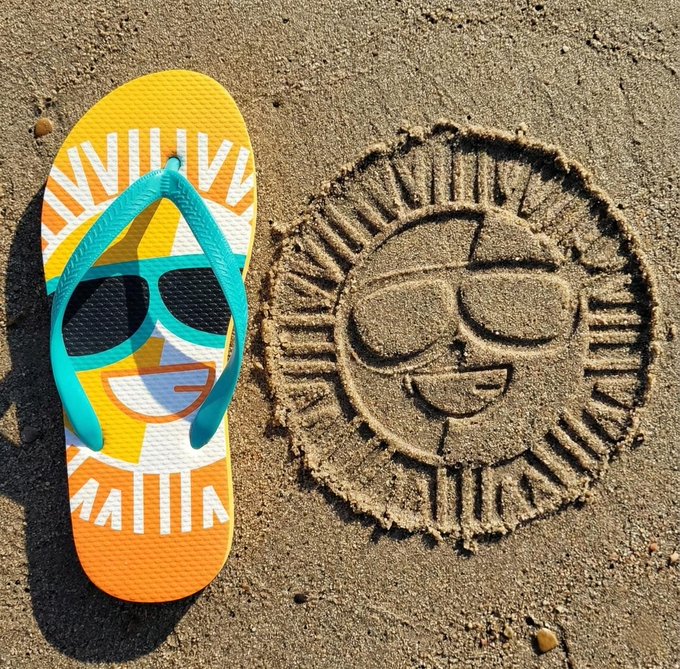 A yellow and orange flip-flop with a sunglasses design lies on sand next to a sand drawing of a smiling sun wearing similar sunglasses.