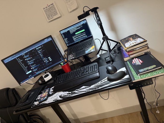 A desk setup featuring a large monitor, a laptop on a stand, a keyboard, ergonomic mouse, books, and a camera mounted above, positioned against a wall with pinned notes.