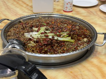 A large metal pan sits on a wooden table, filled with a heavily spiced dish topped with red chili flakes, peppercorns, and sliced scallions.