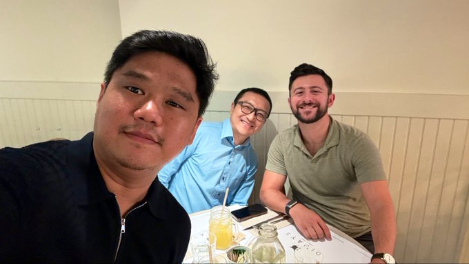 A selfie of three men sitting together at a restaurant table with drinks and a phone, smiling for the camera.